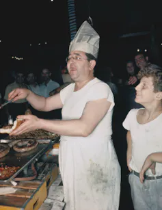 Middle-aged man wearing a greasy, white outfit and a hat made from a newspaper shouts while placing onions, peppers and sausage on a sub roll as a smiling boy looks on.