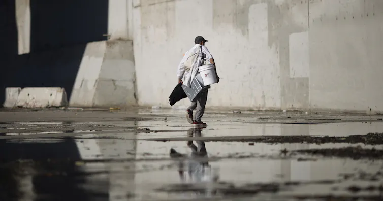 A man carrying several items walks through cement passage with water in it under a bridge