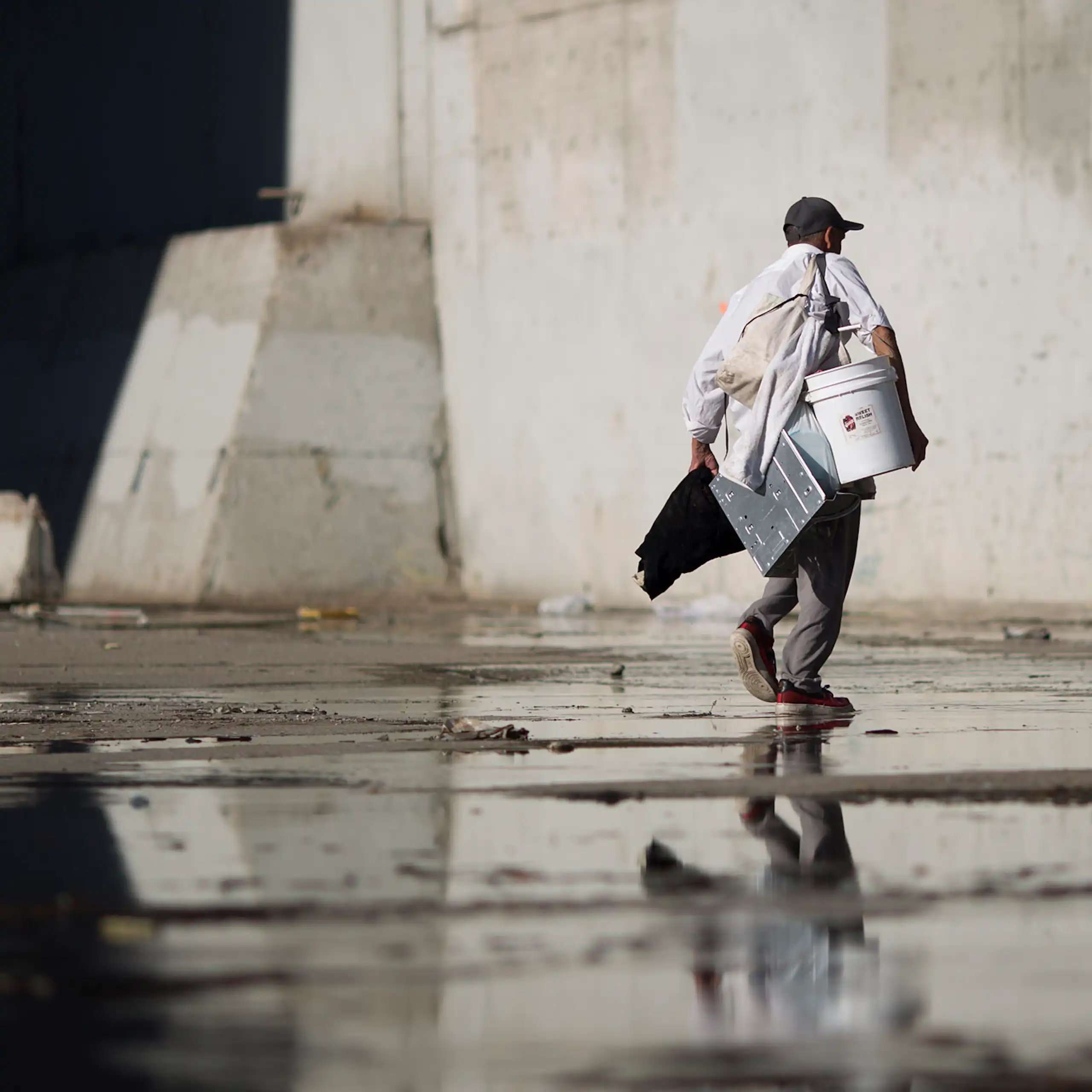 A man carrying several items walks through cement passage with water in it under a bridge