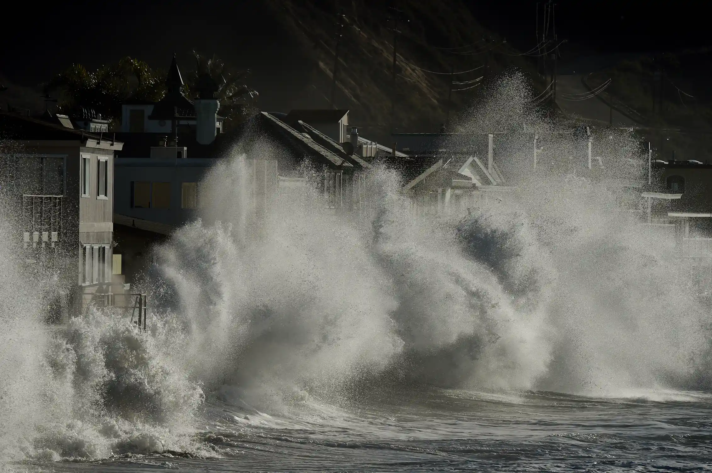 Huge waves crash up against a coastline. Their spray goes as high as the homes that are right next to the water.