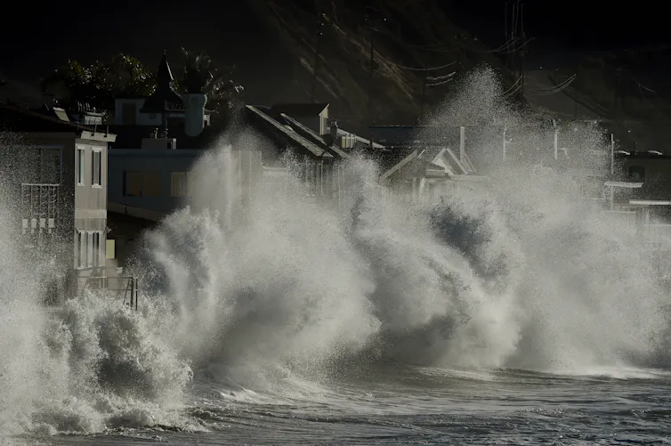 Huge waves crash up against a coastline. Their spray goes as high as the homes that are right next to the water.