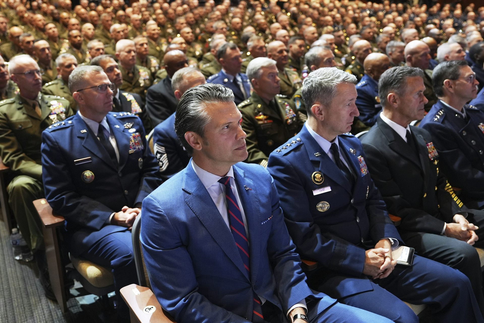 A man in a blue suit sits in an auditorium, with uniformed officers filling the seats beside and behind him.