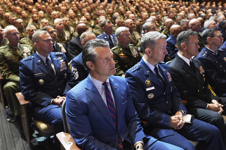 A man in a blue suit sits in an auditorium, with uniformed officers filling the seats beside and behind him.