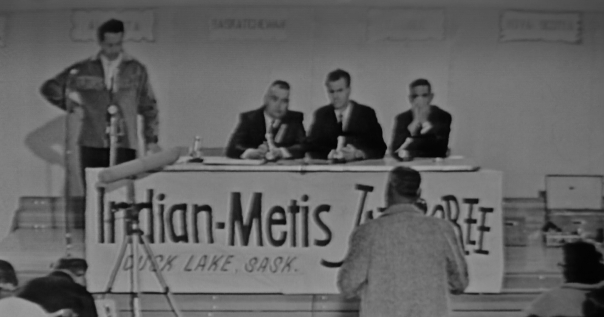 Image shows a banner that says Indian-Metis (the word Jamboree is partly obscured by a person) and Duck Lake, Sk, and people seated at a table in front of spectators.