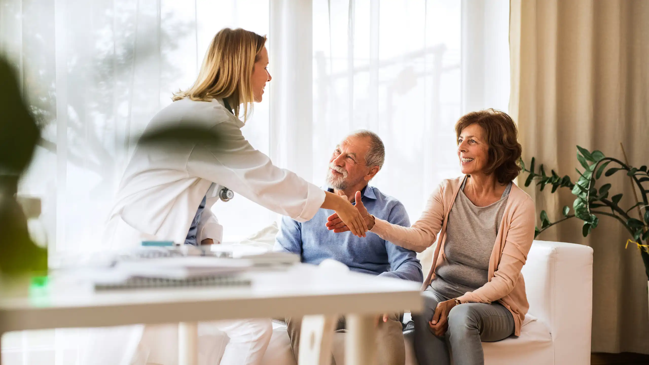 A woman in a white coat shakes hands with another woman seated beside an older man
