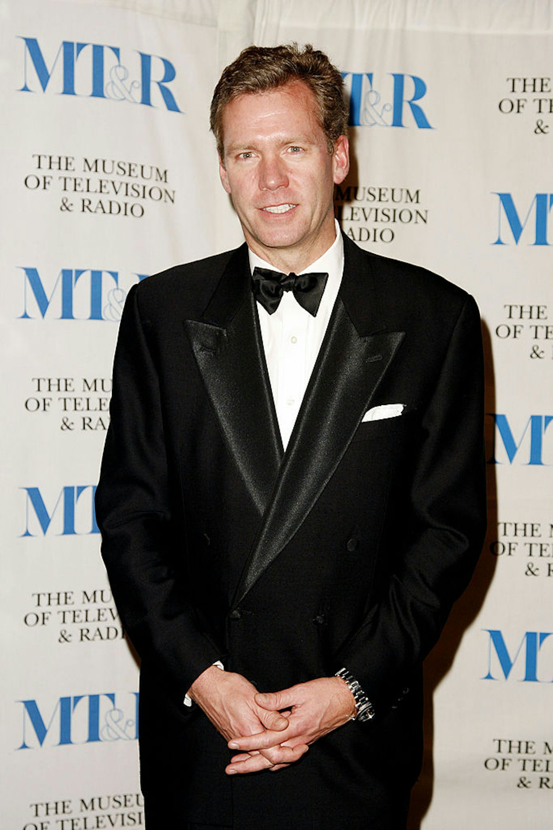 Man wearing a tuxedo poses while clasping his hands at his wasit.