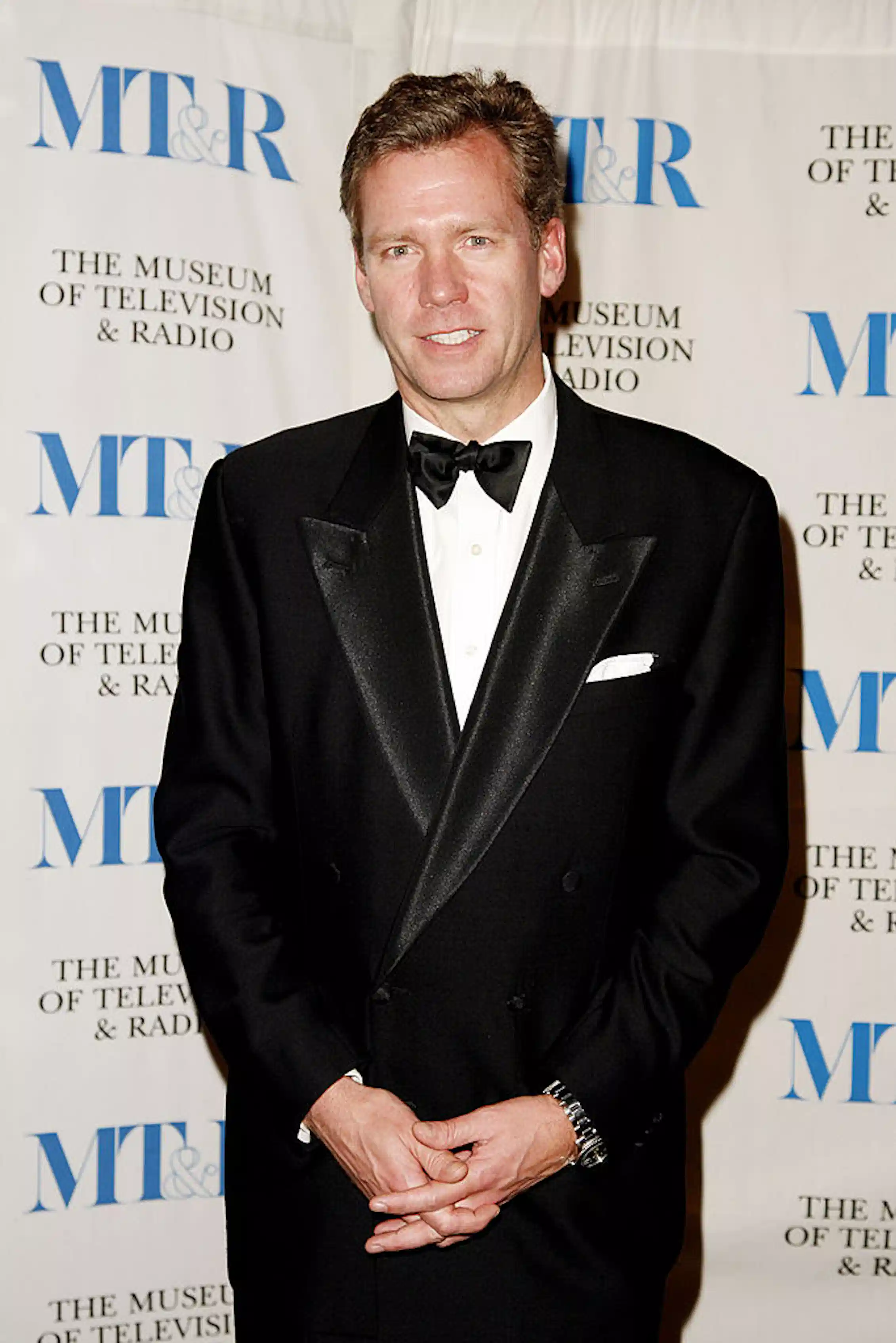 Man wearing a tuxedo poses while clasping his hands at his wasit.