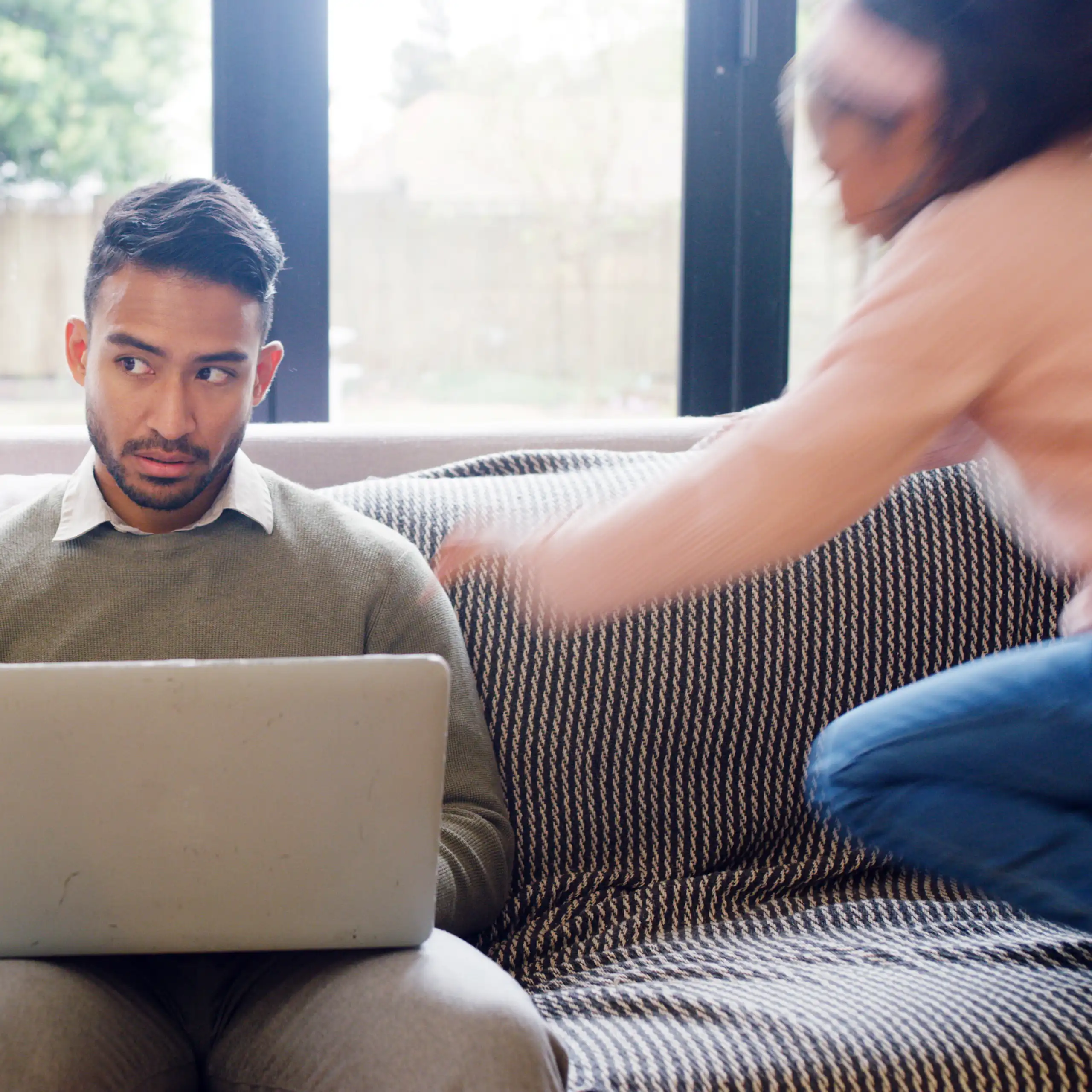 A dad works from home on his laptop sitting on the couch while his two daughters jump and play around him.