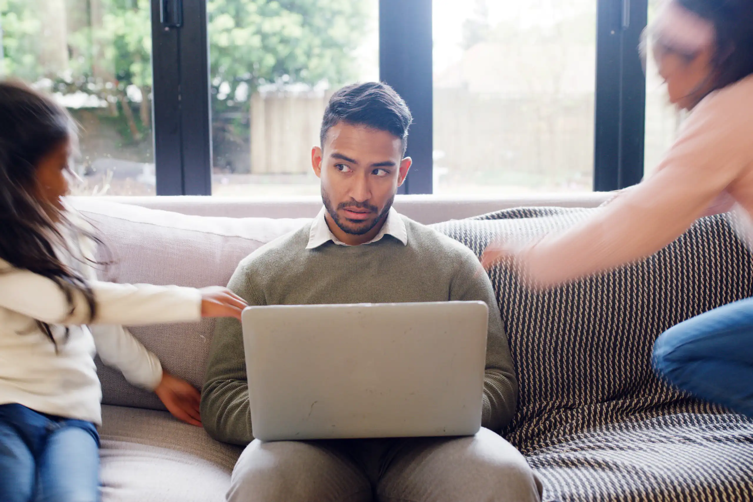A dad works from home on his laptop sitting on the couch while his two daughters jump and play around him.