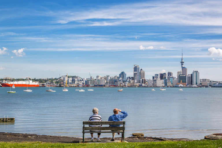 Couple on park bench looking across water at Auckland CBD on clear day