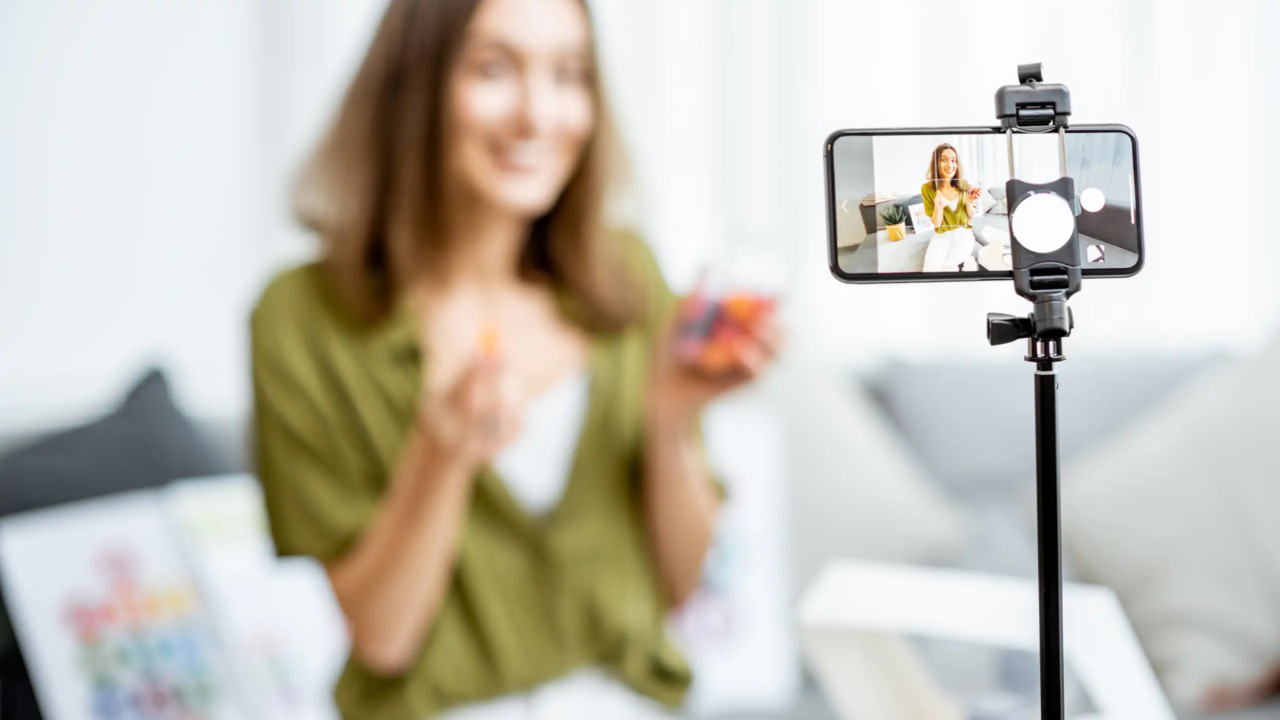 Female influencer sitting on couch holding supplements looking at smartphone on stand