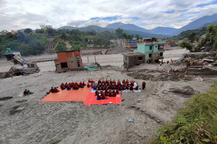 Buddhist monks sit on red tarps in a muddy expanse conducting death rituals