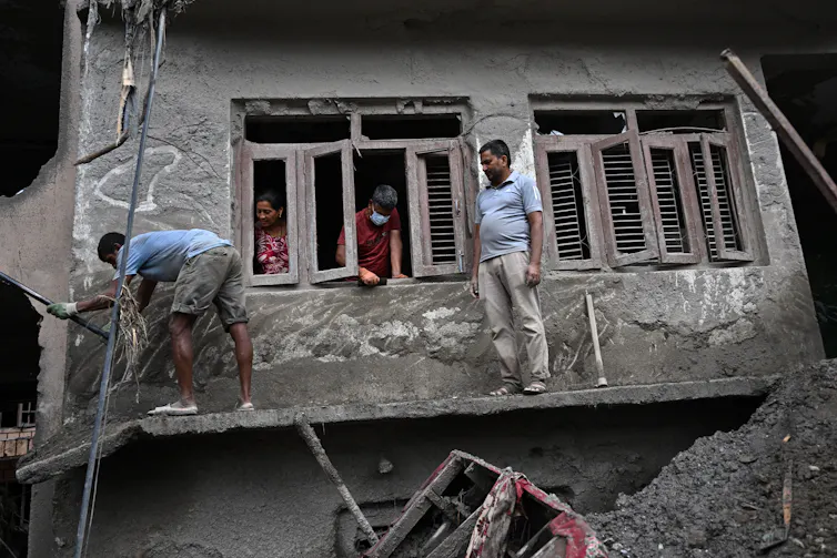 People stand on a board to reach second-floor windows of a home. The mud largely covers the first floor.
