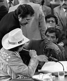 Two men conferring at a table at a political event with people behind them.