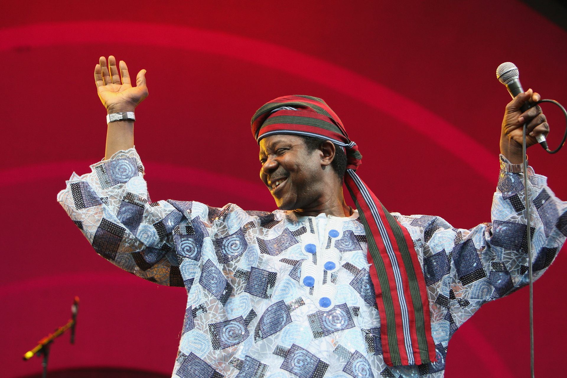 A man in African style loose shirt and headscarf, holding a microphone with hands raised