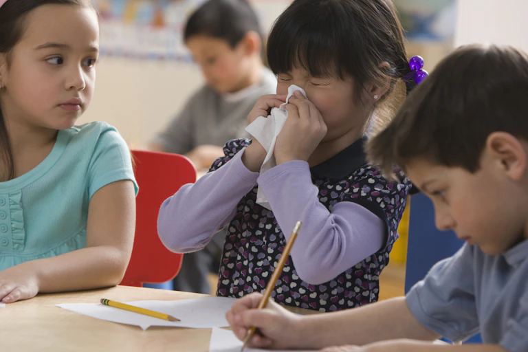 Three elementary-aged children working at a table in school, the one in the middle is blowing her nose