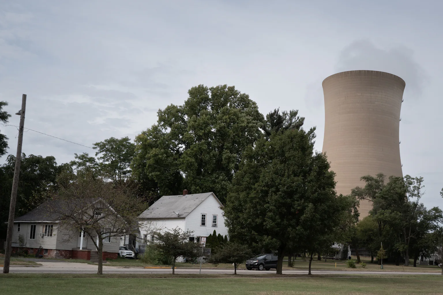 A cooling tower looms above a house and grove of trees.