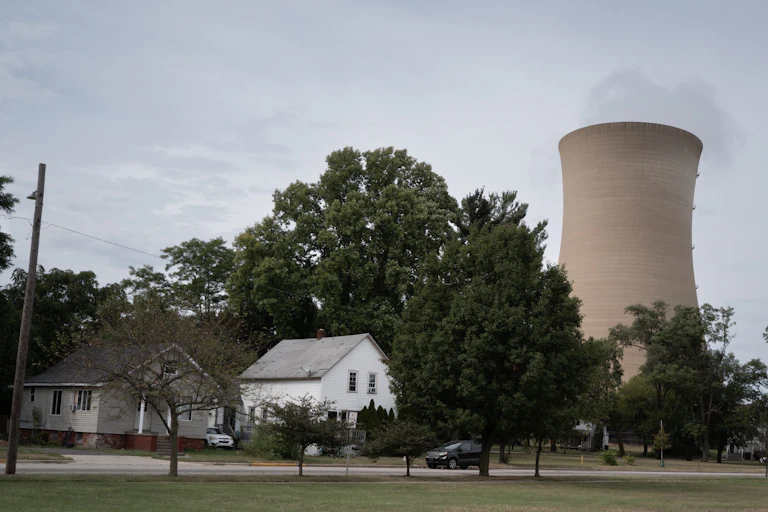 A cooling tower looms above a house and grove of trees.