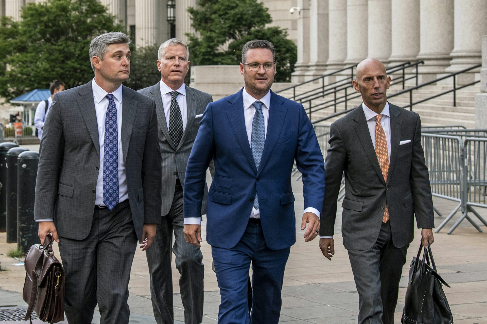 Four men in suits walk along a sidewalk outside a courthouse, two carrying briefcases.