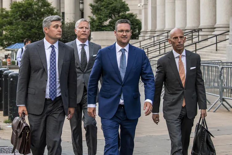 Four men in suits walk along a sidewalk outside a courthouse, two carrying briefcases.