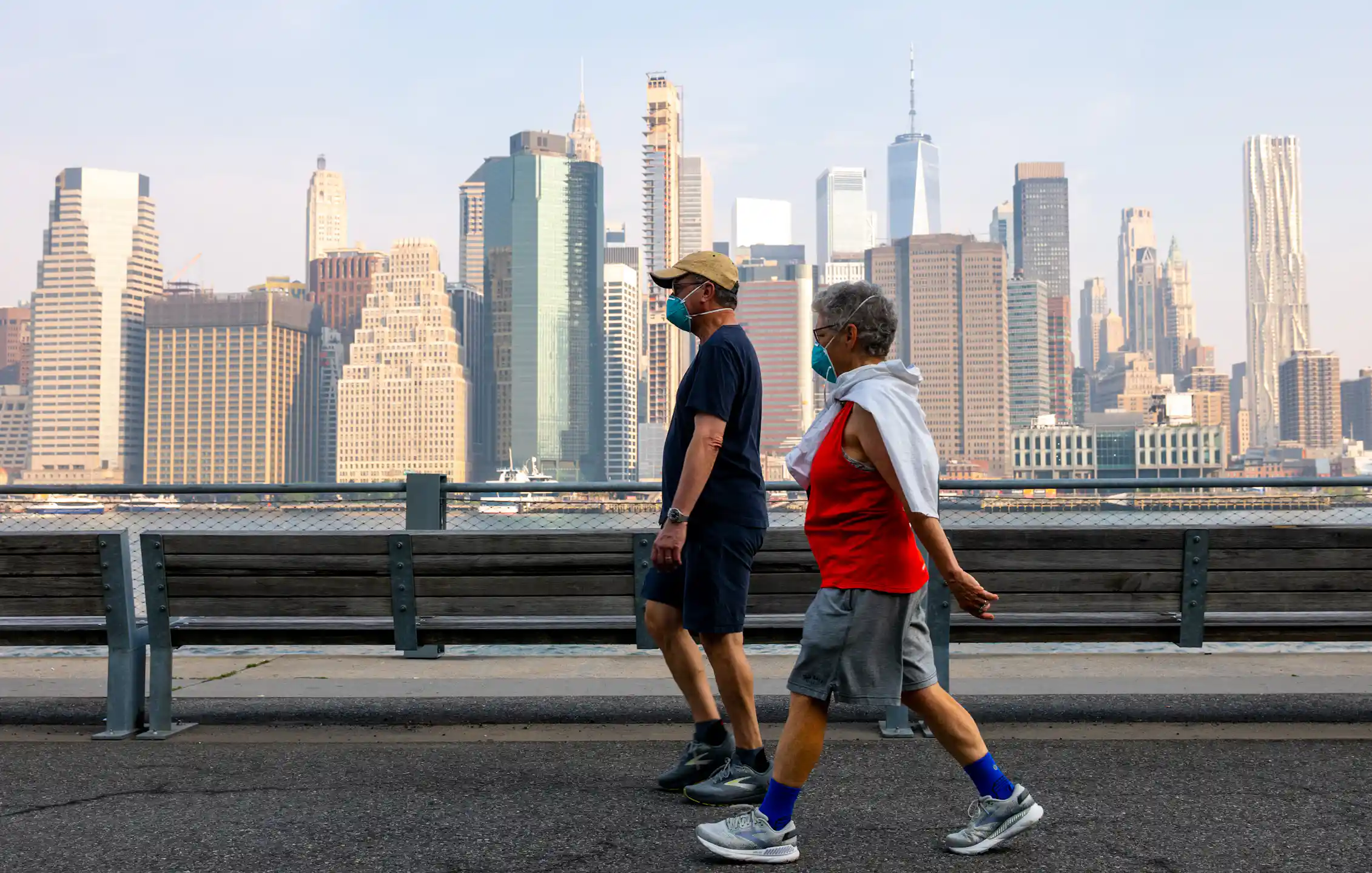 Two people wear masks over their nose and mouth while they walk along a path with a cityscape visible in the background.
