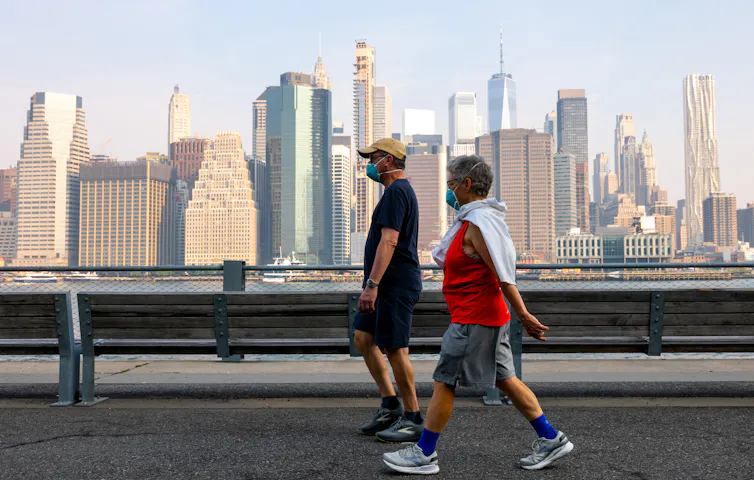 Two people wear masks over their nose and mouth while they walk along a path with a cityscape visible in the background.