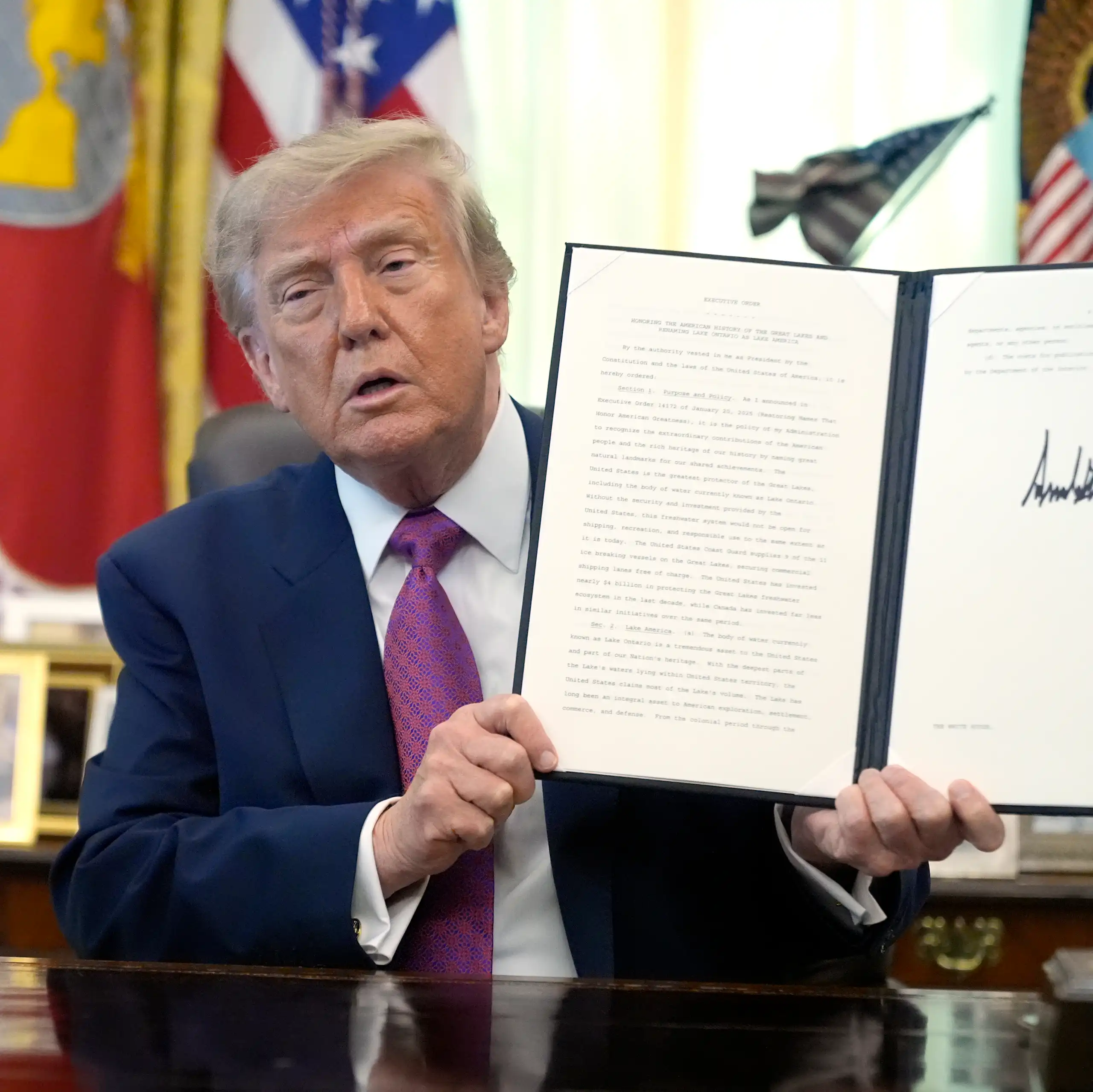 A man in a suit sits at a desk in front of flags and holds up an open folder with a signed document inside.