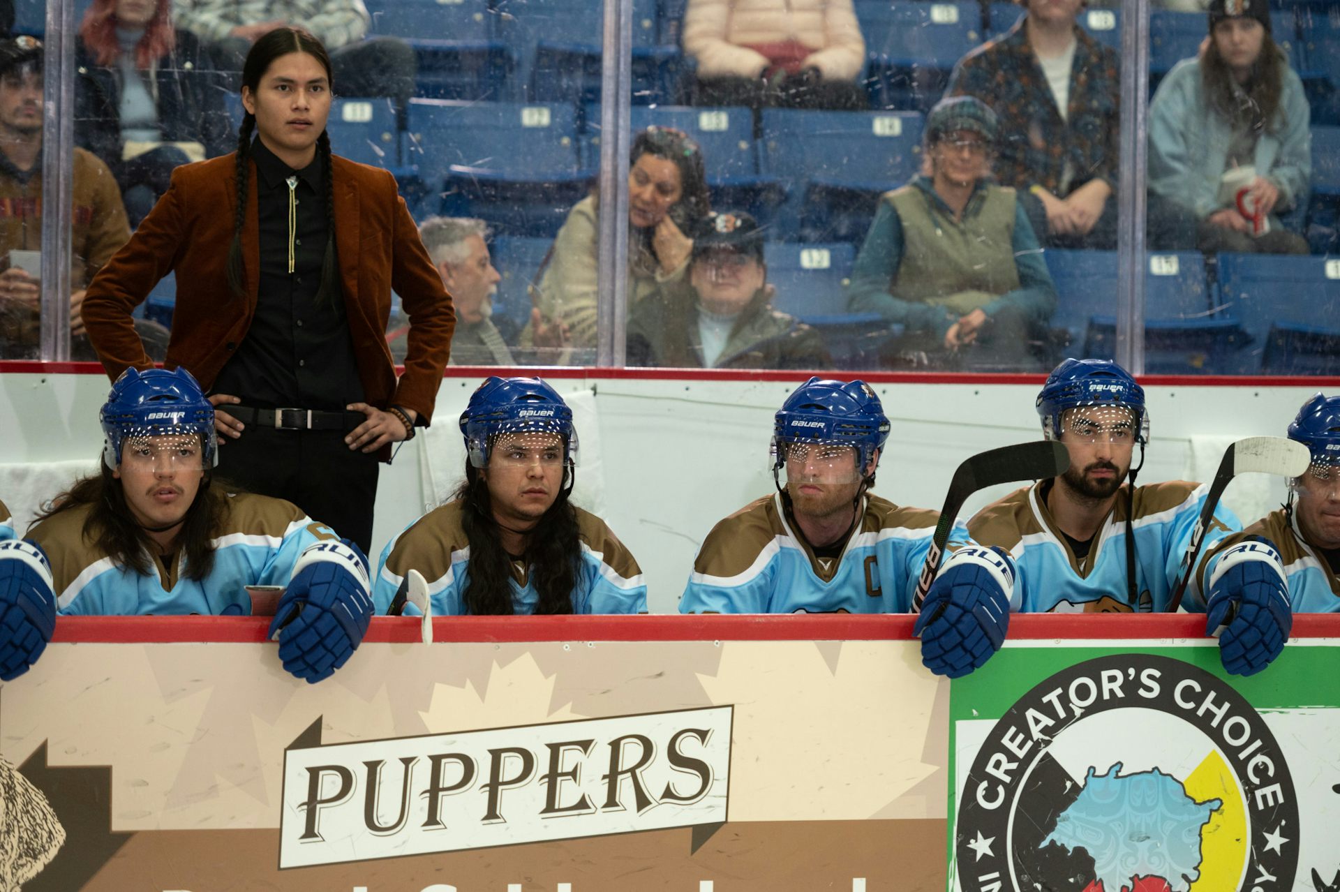A Cree man, with his hair in two long braids and wearing a suit, stands behind a bench of hockey players