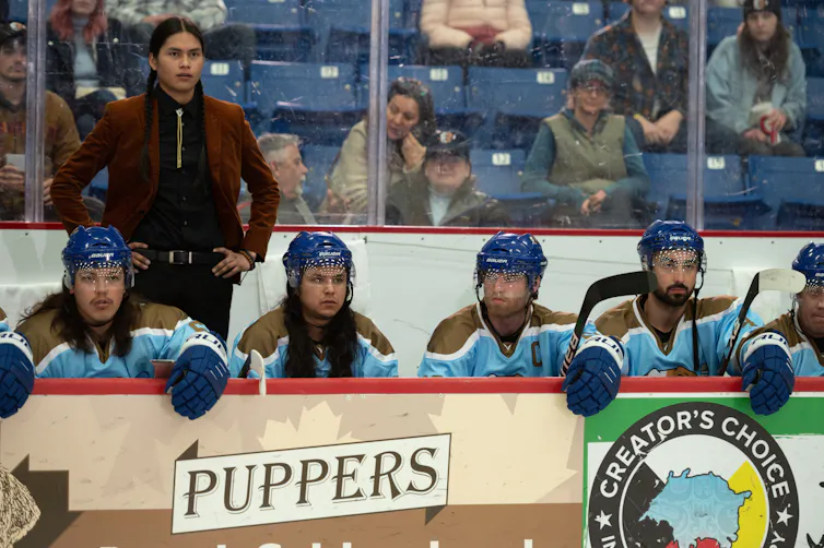 A Cree man, with his hair in two long braids and wearing a suit, stands behind a bench of hockey players