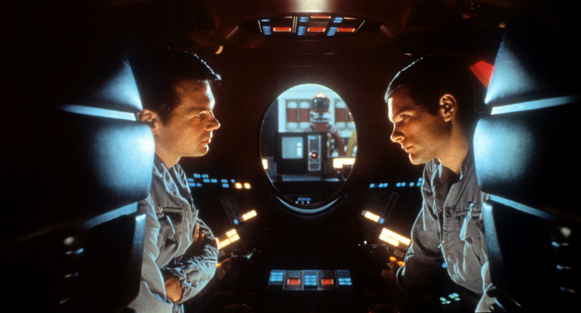 dois homens frente a frente dentro de um espaço apertado, repleto de botões iluminados e com uma janela oval