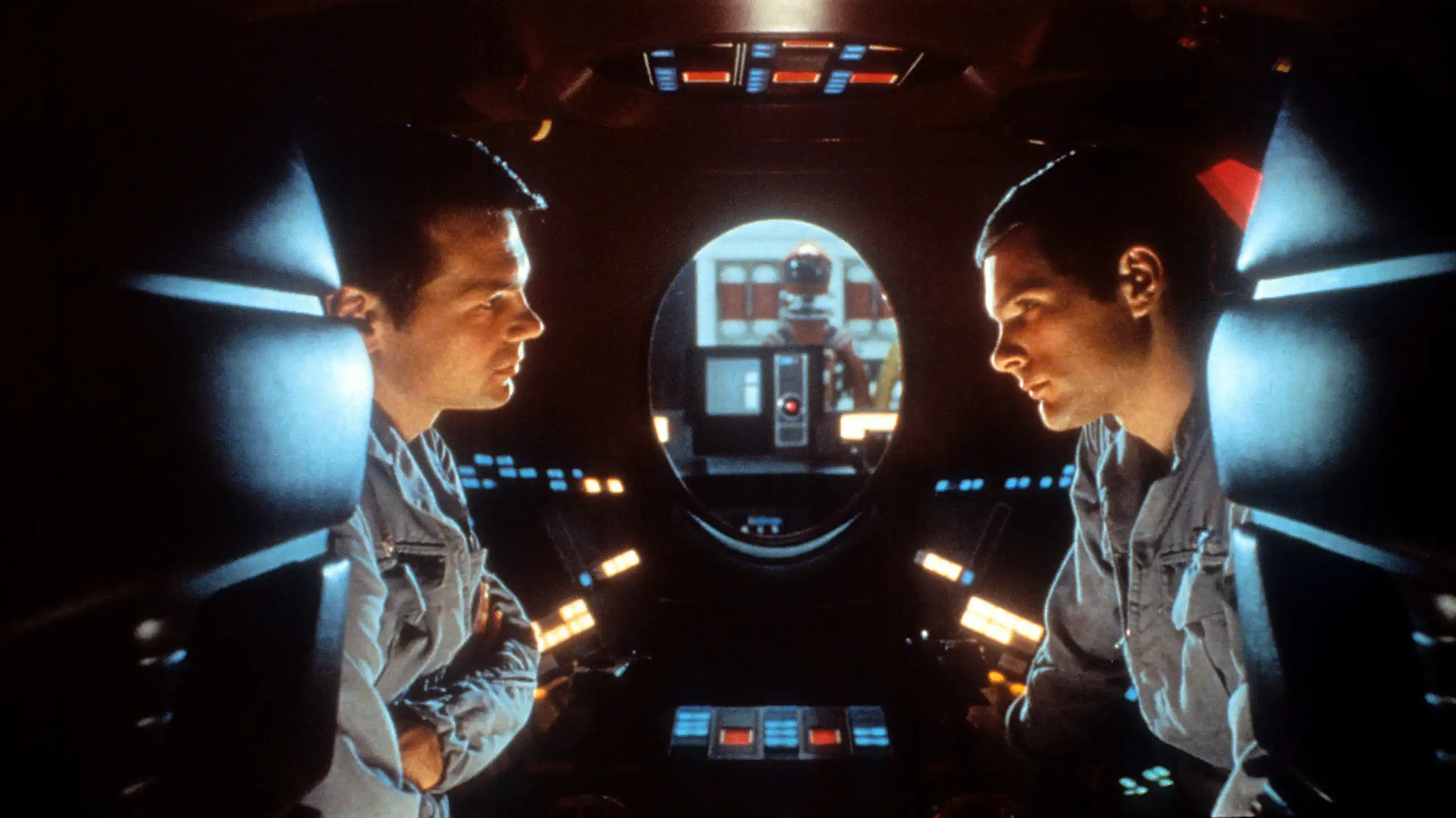 dois homens frente a frente dentro de um espaço apertado, repleto de botões iluminados e com uma janela oval