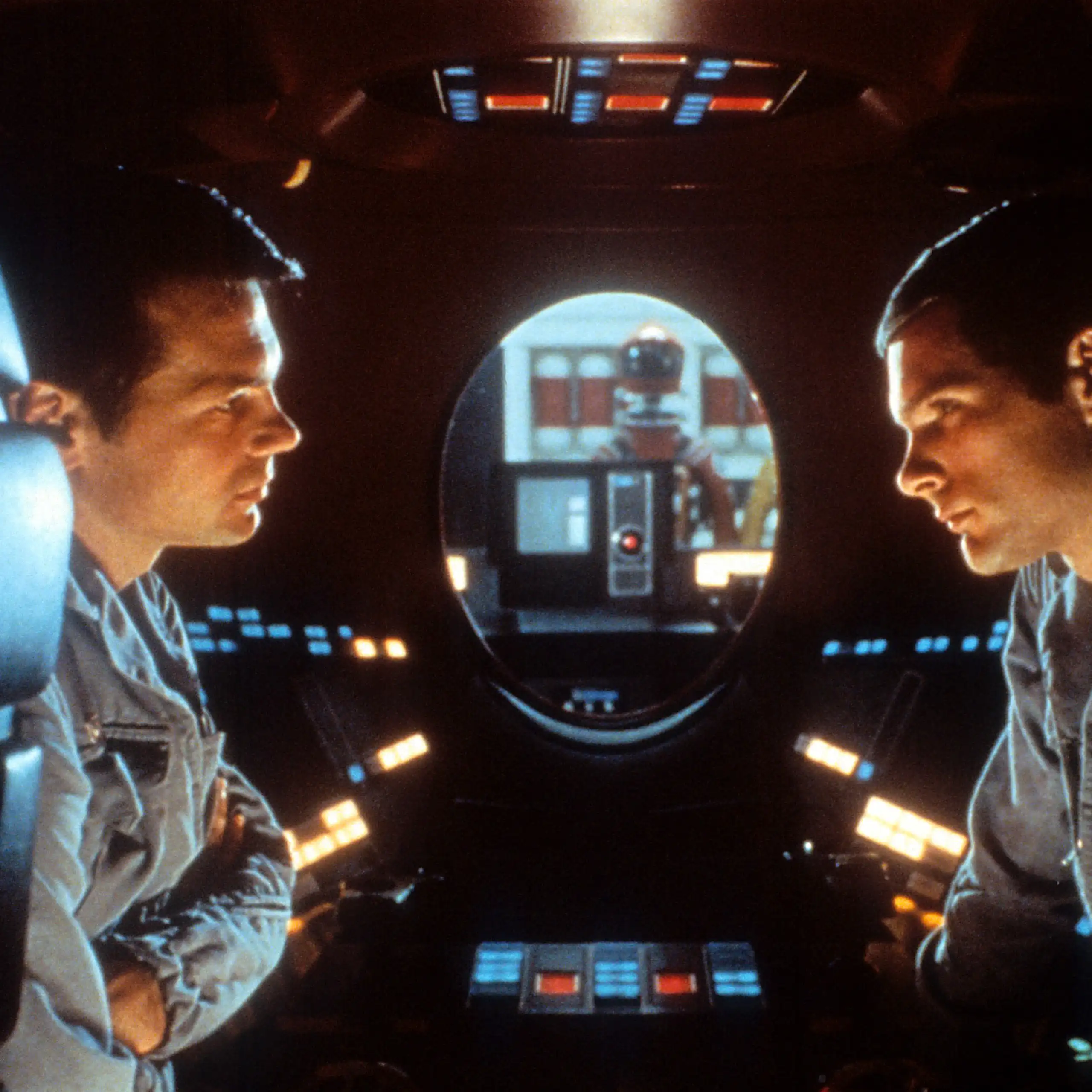 dois homens frente a frente dentro de um espaço apertado, repleto de botões iluminados e com uma janela oval