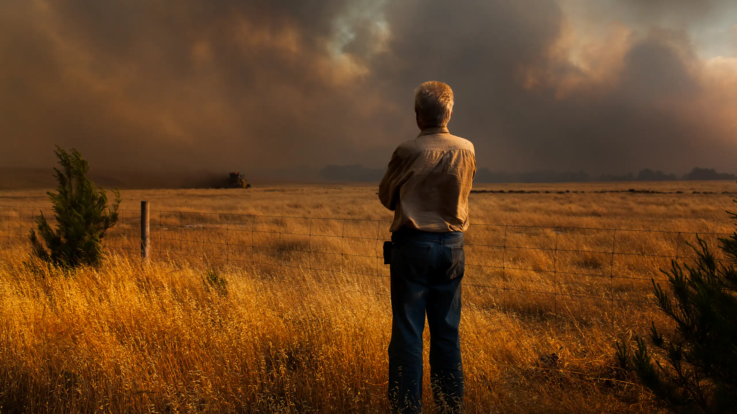 A man standing in a paddock, watching a bushfire burn in the distance.