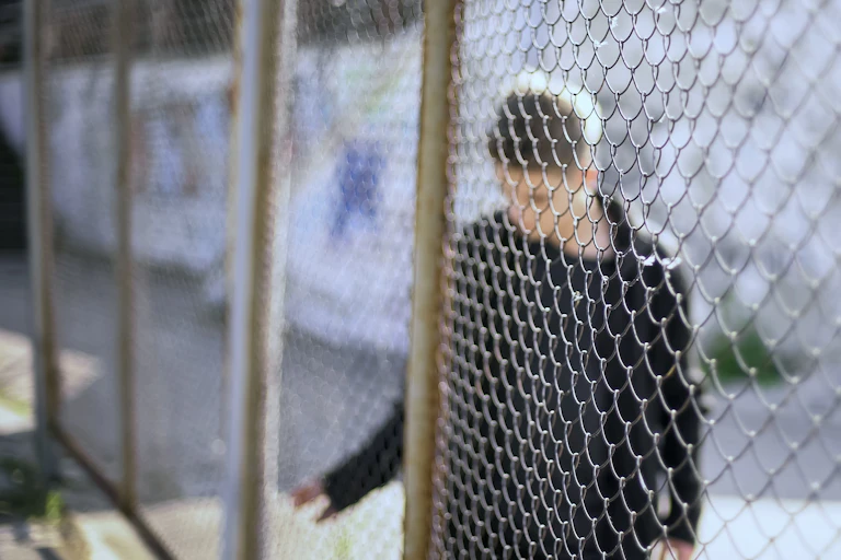 A young man with a blurred out face is seen behind a wire fence.
