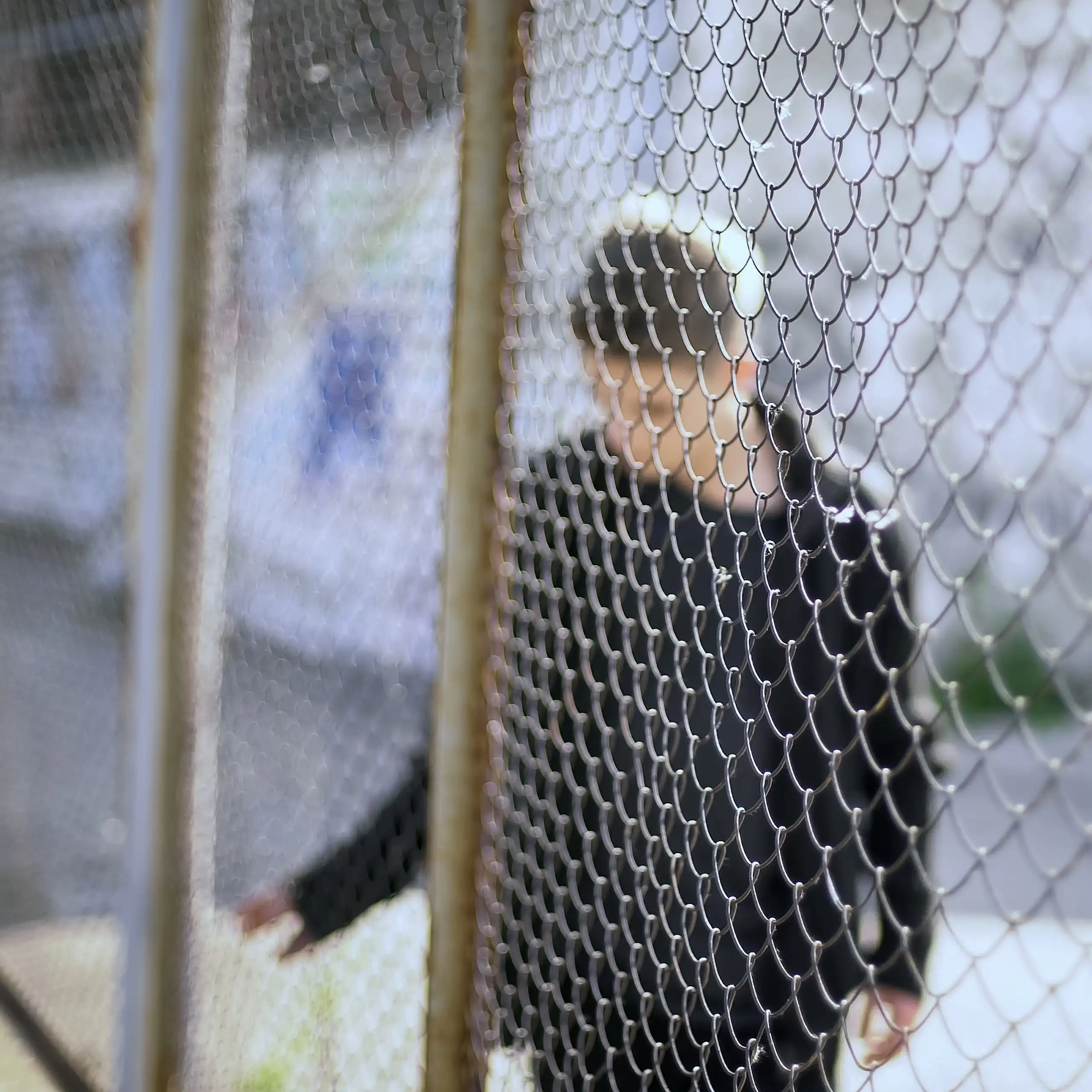 A young man with a blurred out face is seen behind a wire fence.