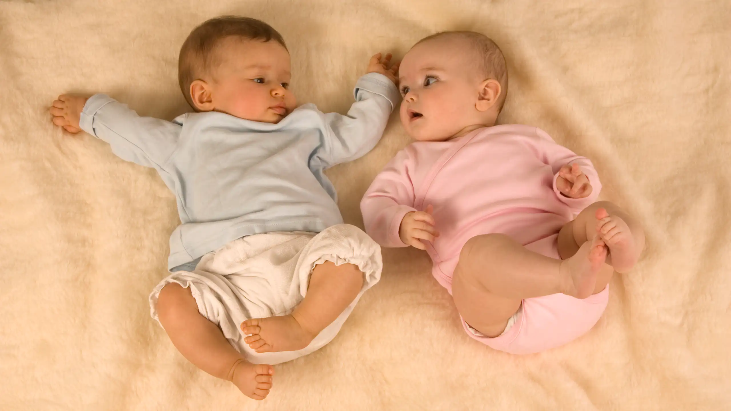 A baby in blue laying beside a baby in pink on a bed, both looking at each other.