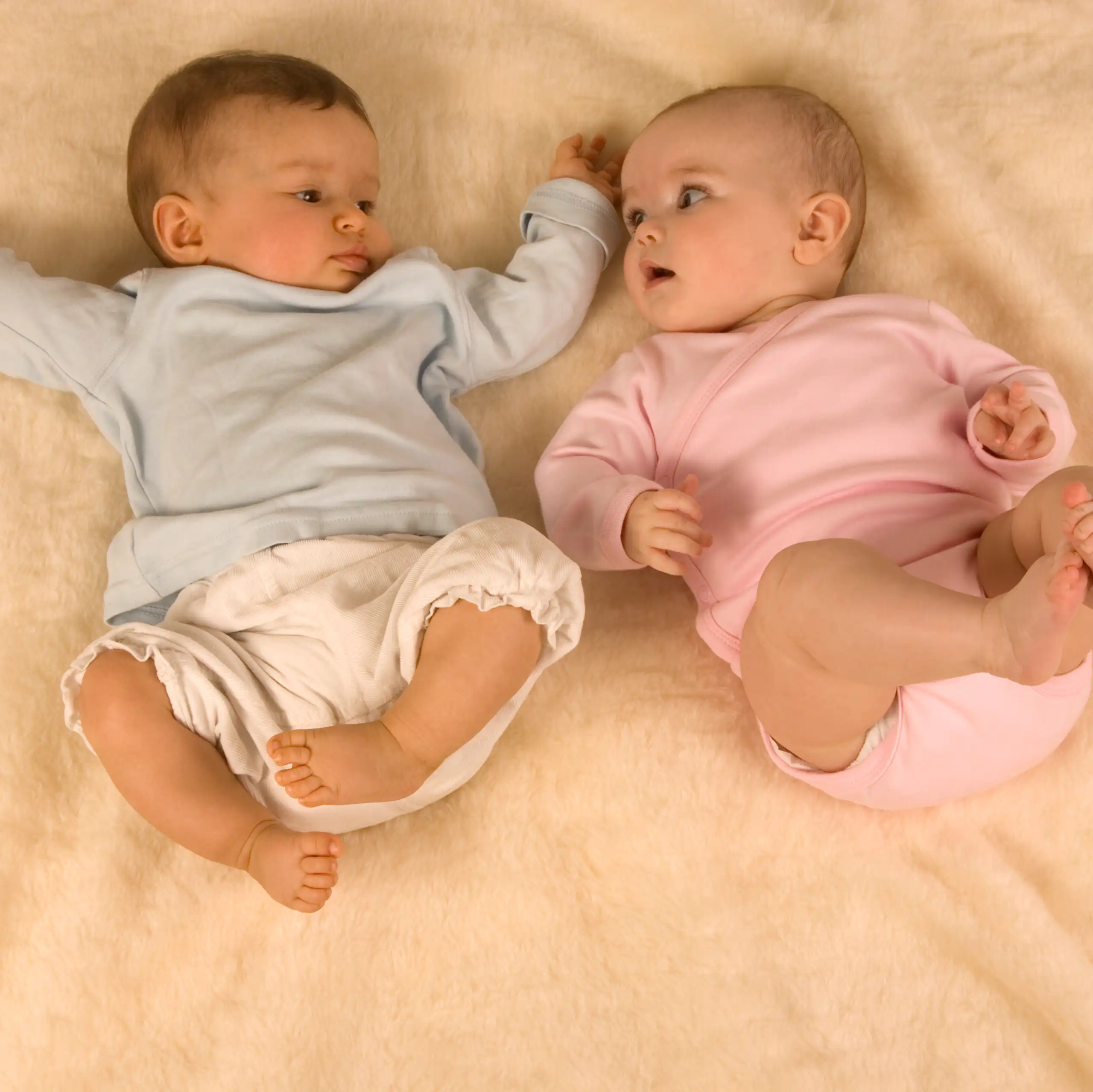 A baby in blue laying beside a baby in pink on a bed, both looking at each other.
