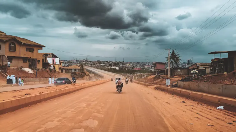 A dirt road with a motorcycle on it, heavy clouds in the sky and houses and buildings in the distance.