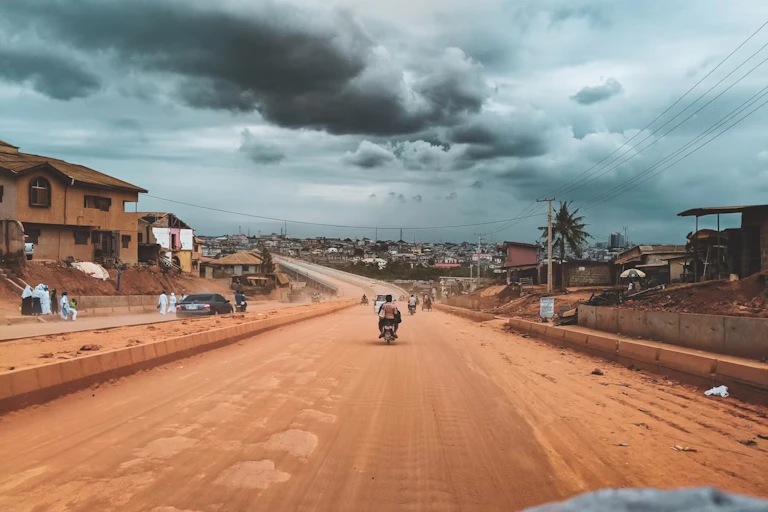 A dirt road with a motorcycle on it, heavy clouds in the sky and houses and buildings in the distance.
