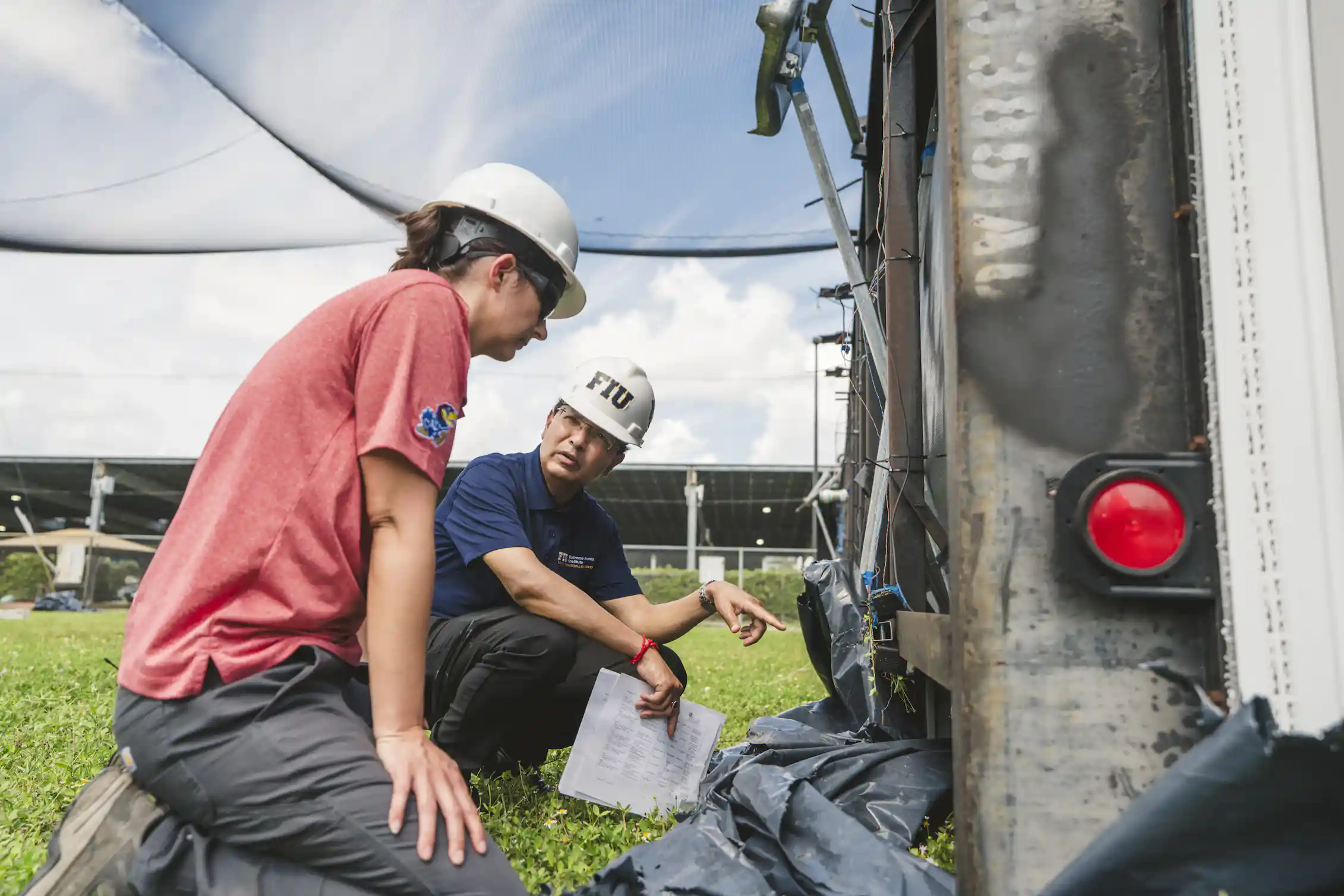 Two people in hard hats examine the bottom of a tipped-over mobile home.