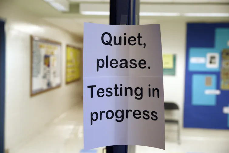 A white sign with black font says 'Quiet, please. Testing in progress,' with an empty school hallway behind it.