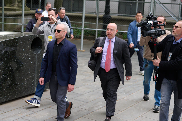 A man dressed in a dark suit and dress shirt walks while another man in a suit trails him and photographers shoot his picture.