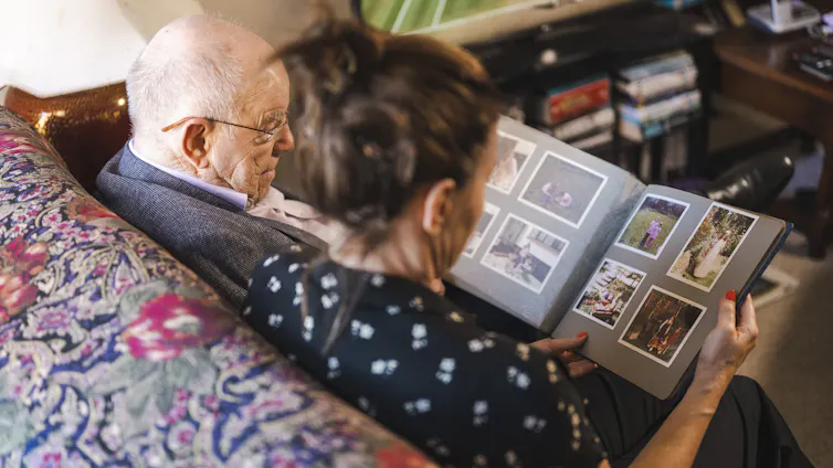 An older man sits on a couch with a younger woman, looking at a photo album together.