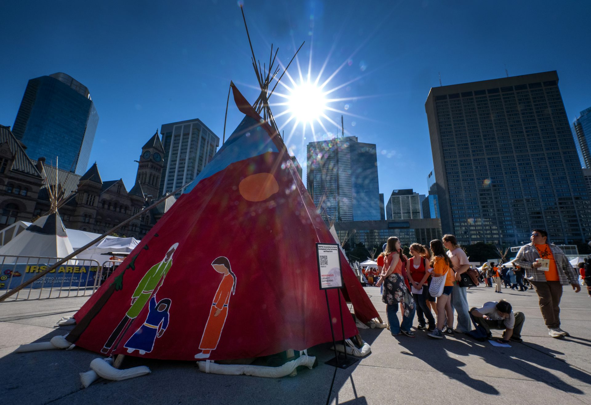 A bright sun in the sky over a teepee and students in orange shirts are standing next to it. 