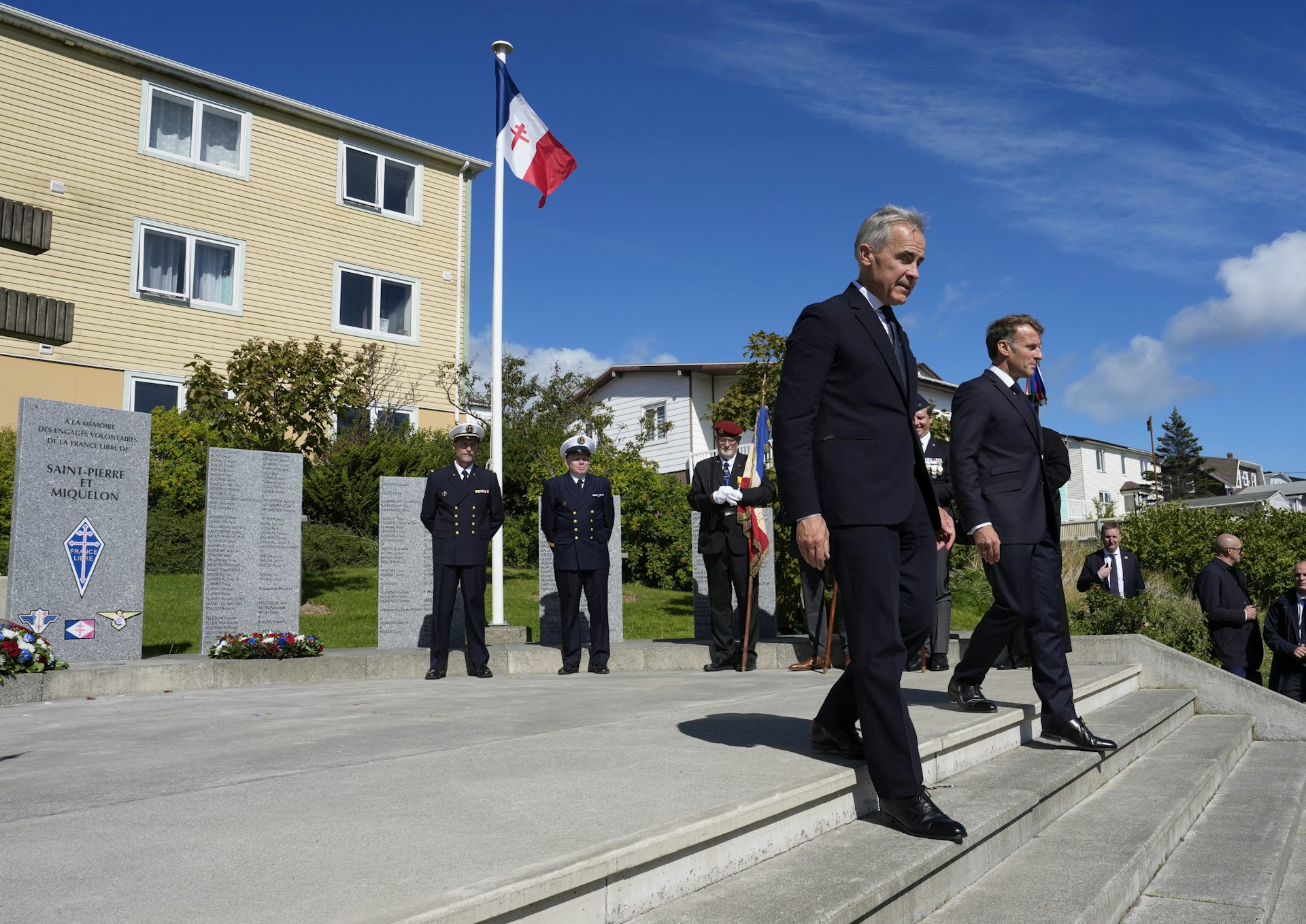 Two men in suits walk down a short flight of stairs as a French flag flies behind them on a sunny day.