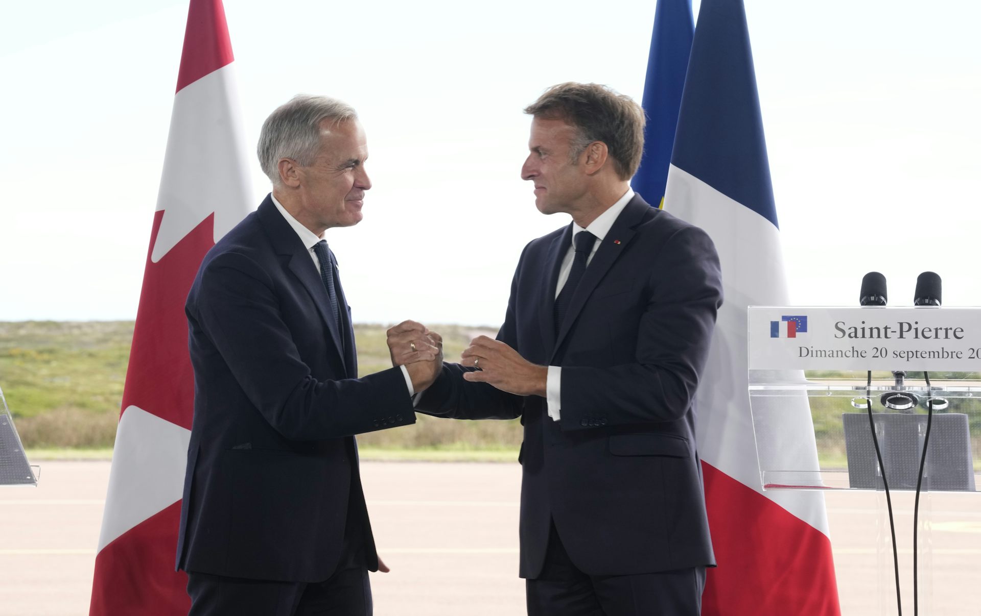 A man with short grey hair and a man with short dark hair clasp hands standing front of the Canadian and French flags on a sunny day.