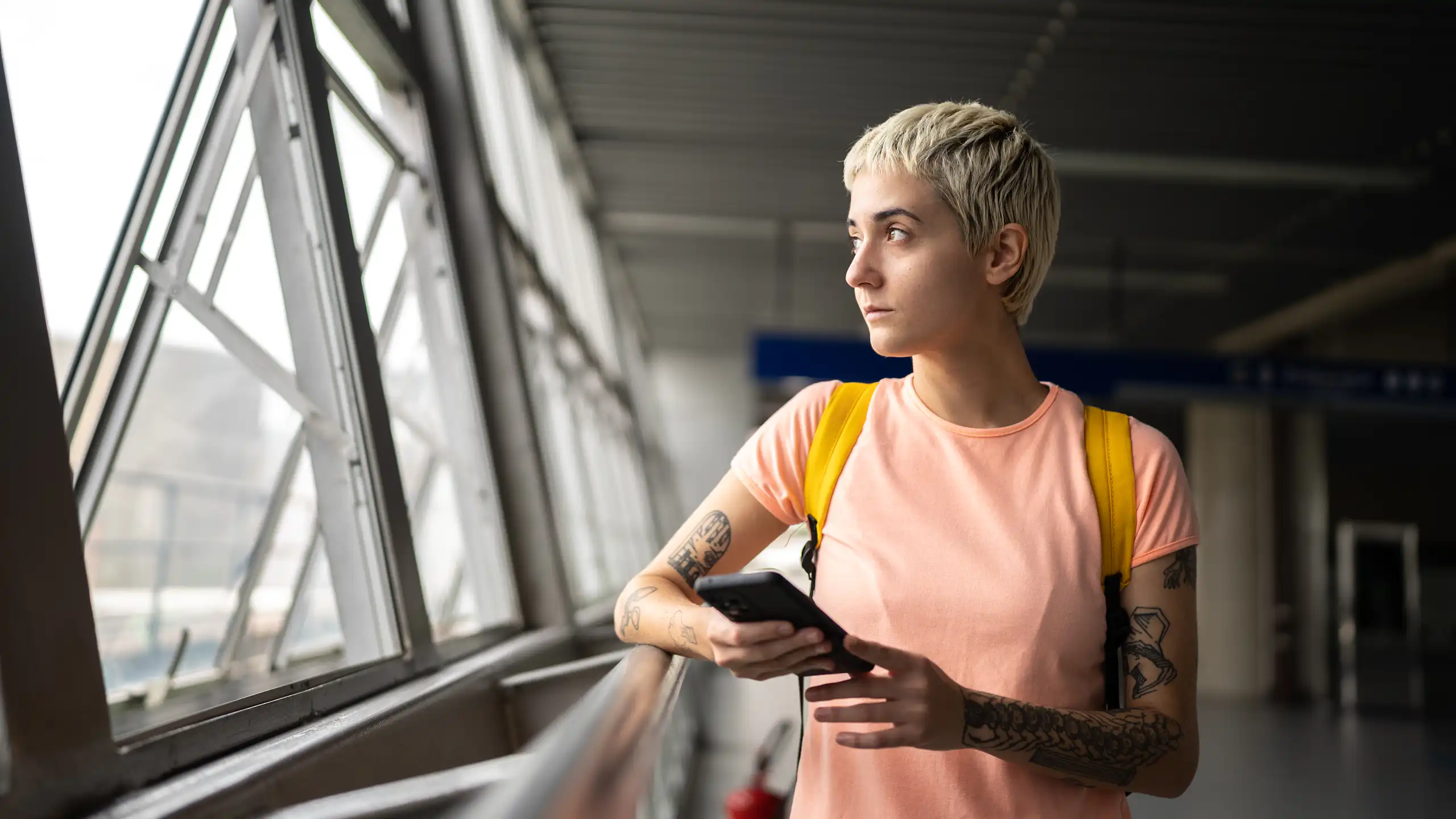 Young person with tattoos and short hair looks out window.