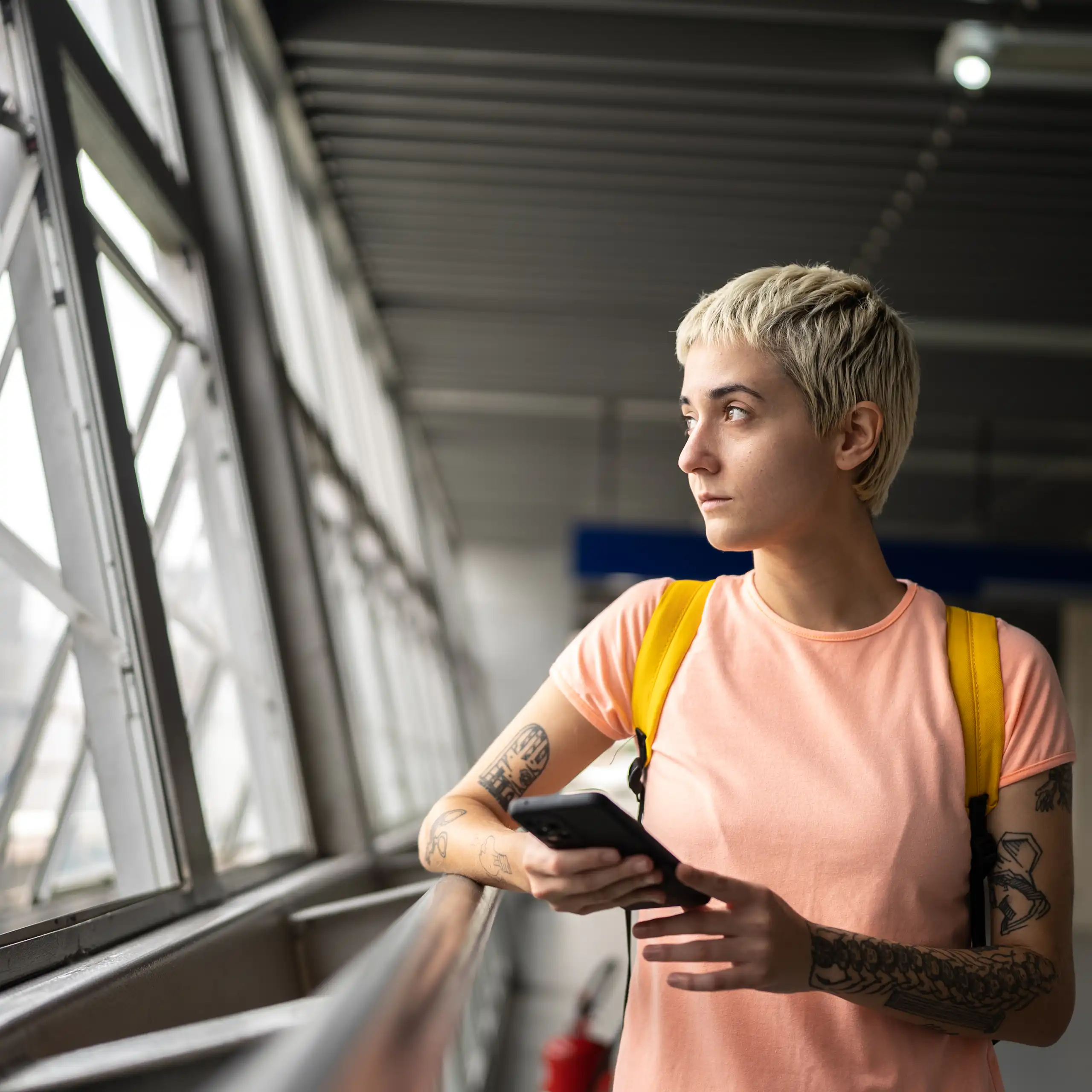 Young person with tattoos and short hair looks out window.