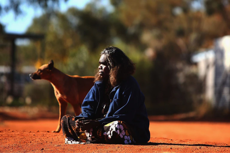 An Indigenous woman sits on red dirt with a dog