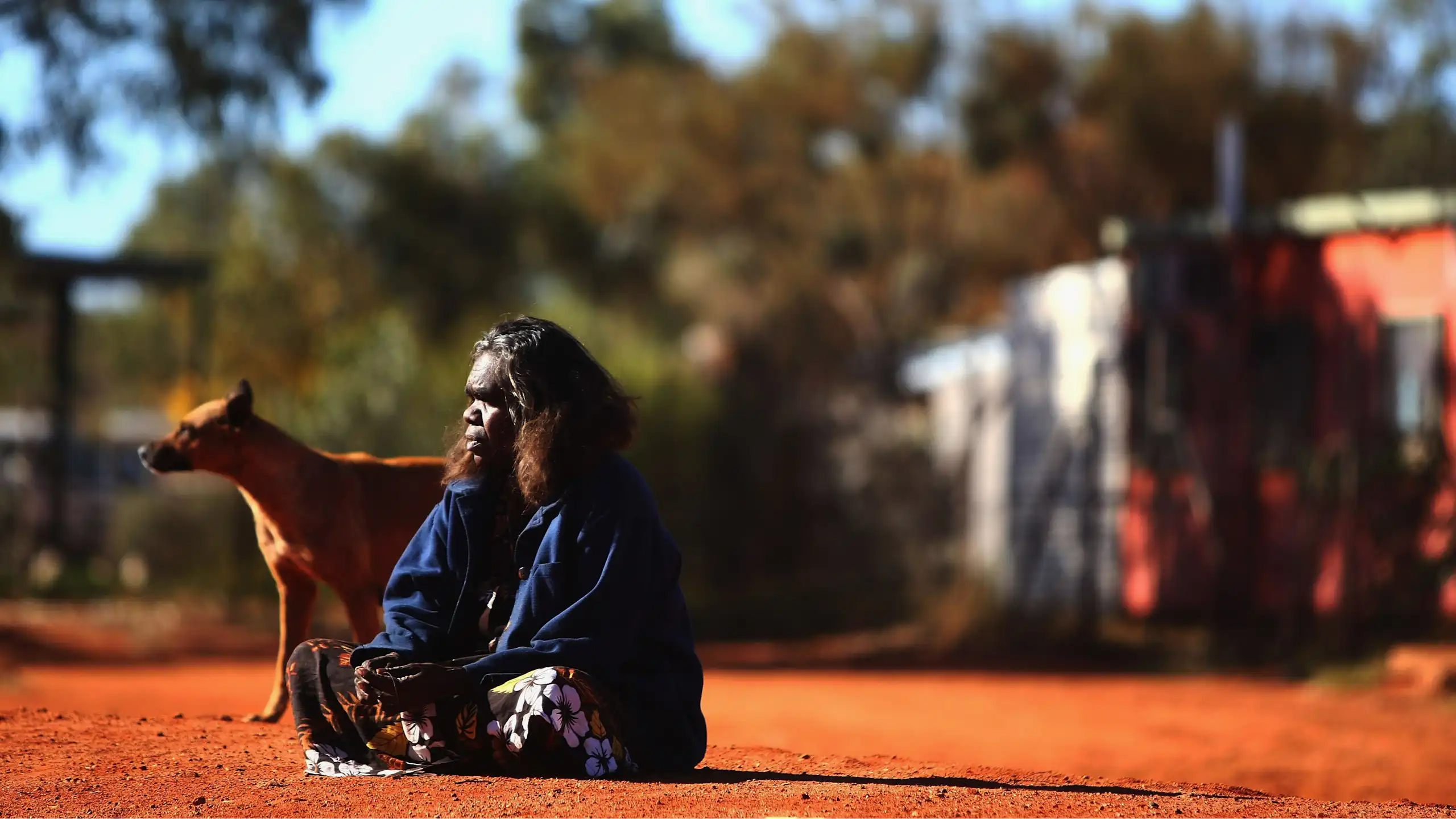 An Indigenous woman sits on red dirt with a dog